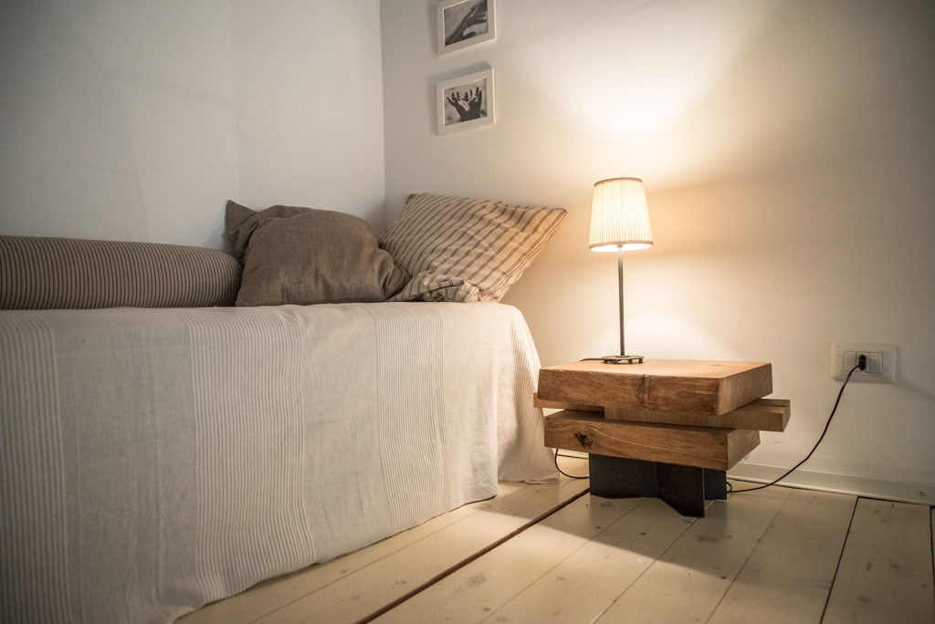 Via delle Orfane by CON3STUDIO - Desk Lamp On Wooden Table Beside Grey Bed Added Near Framed Pictures On Wall